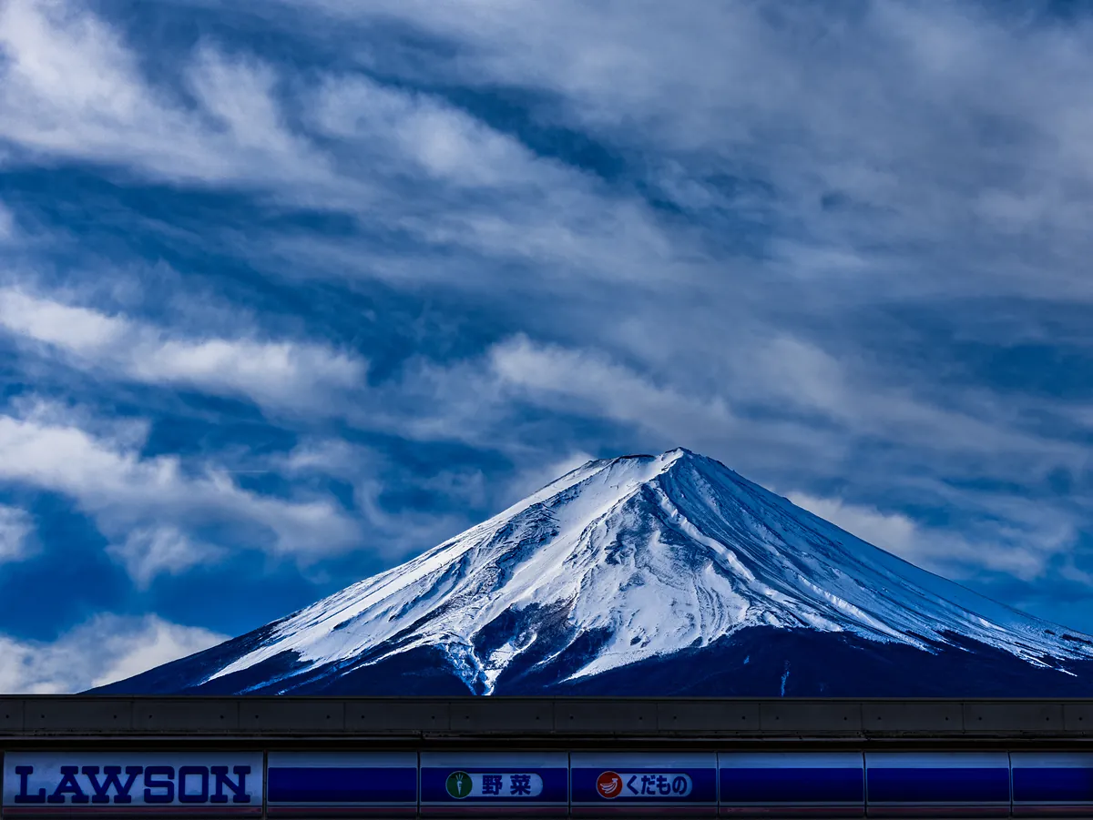 Mount Fuji's snow-covered peak under streaked cloud, rising above the blue roofline of a Lawson convenience store.