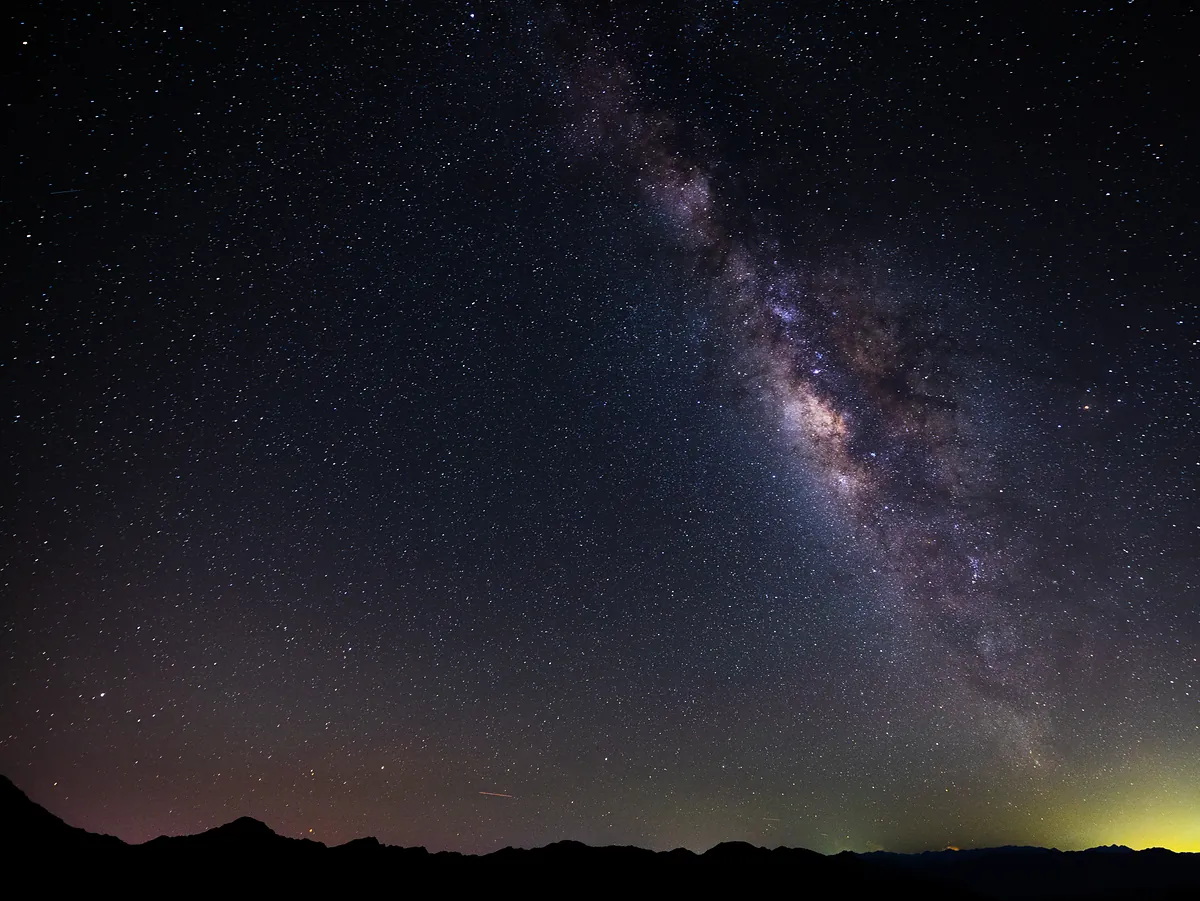 The Milky Way arcs over a dark mountain ridge, the sky dense with stars and a meteor trail at left.