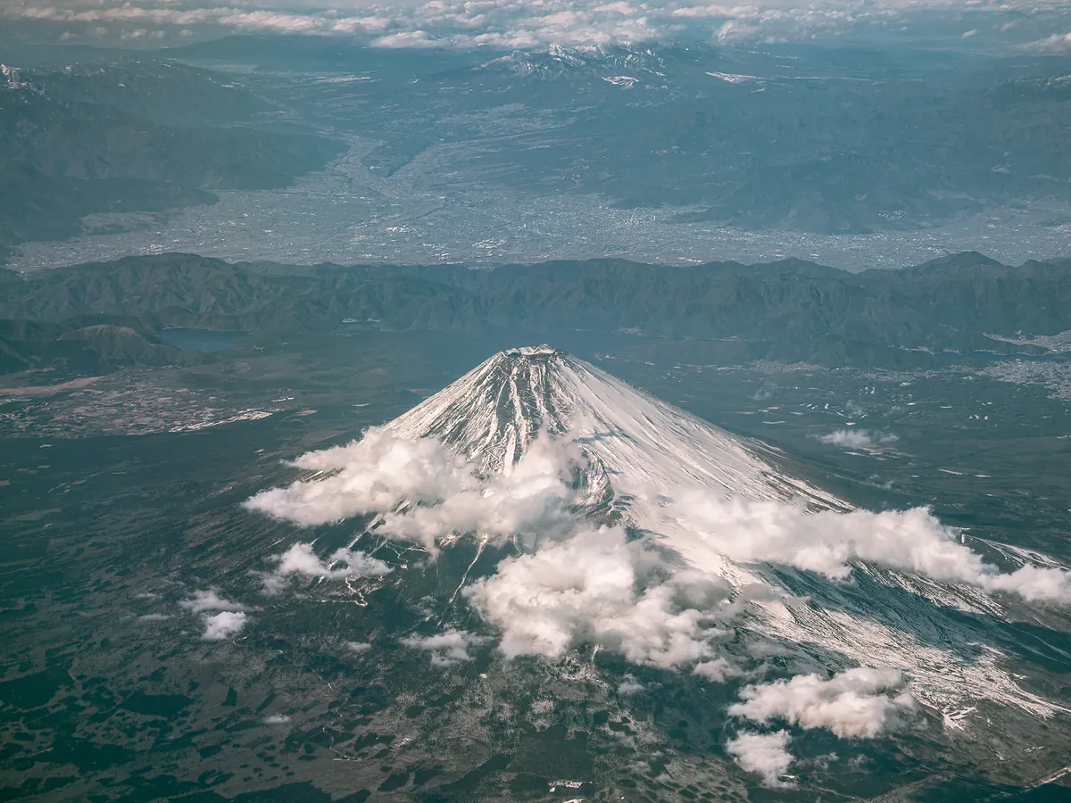 Aerial view of snow-capped Mount Fuji with small clouds on its slopes and valleys beyond.