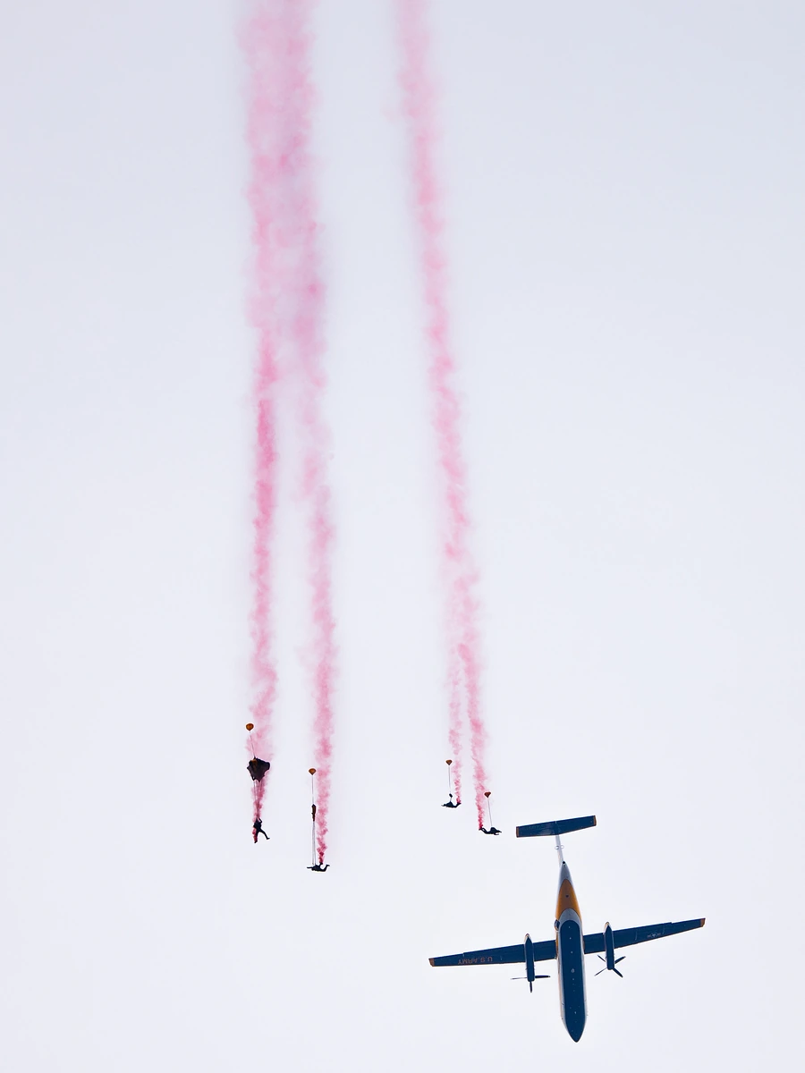 Parachutists trailing pink smoke drift down beside a U.S. Navy aircraft against a pale sky.