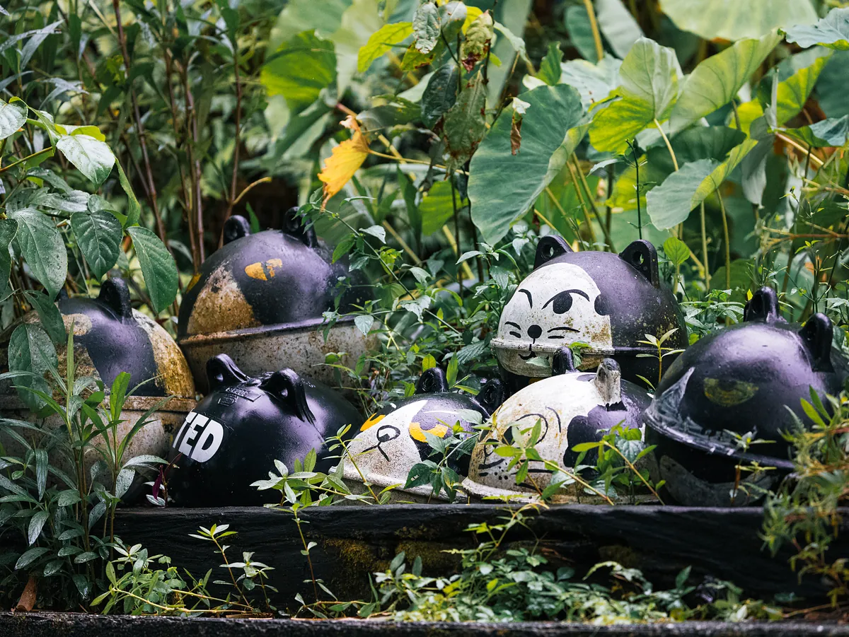 Painted cat-faced figures half hidden among broad tropical leaves.