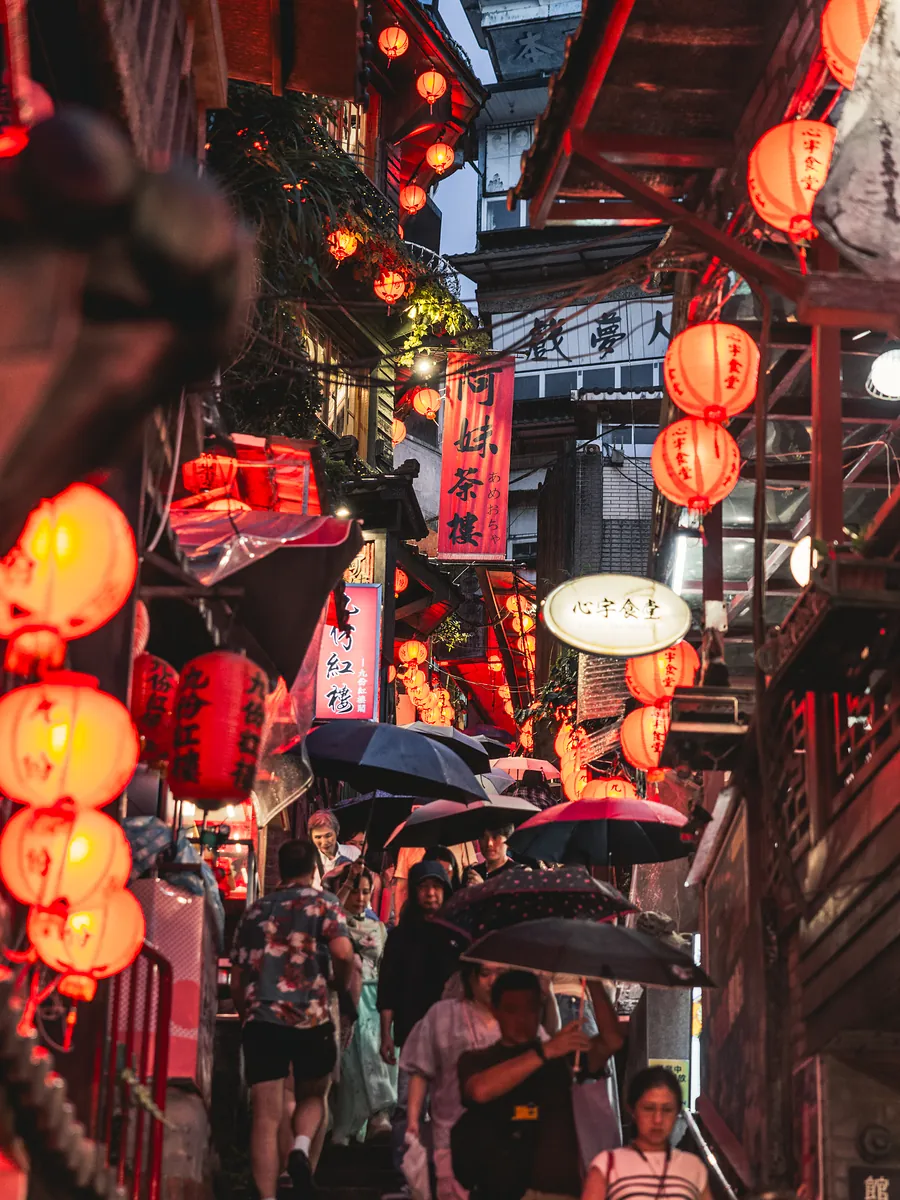 A narrow stepped street hung with red lanterns, crowded with people under umbrellas.