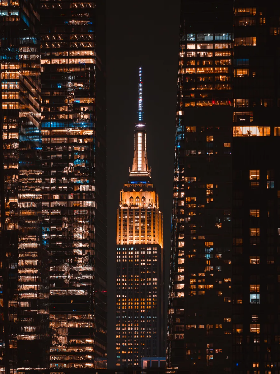 The Empire State Building glowing gold at night, framed between two dark glass towers full of lit windows.
