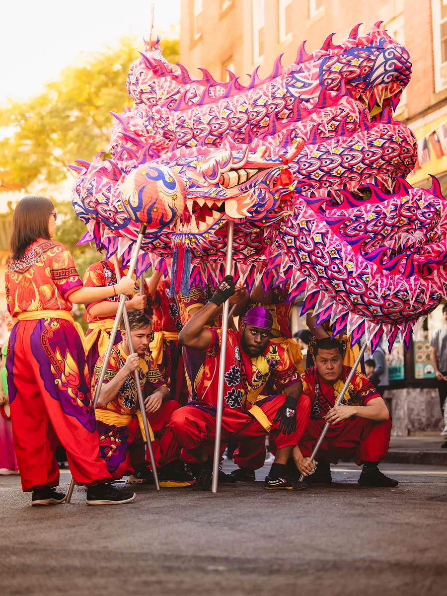 Dancers in red and gold crouch under a huge pink and purple dragon on a city street.