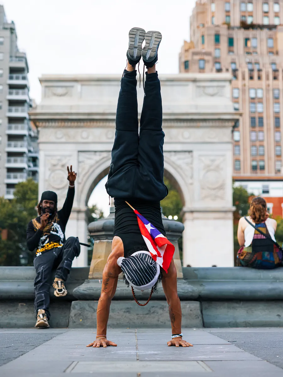 A performer holds a handstand in front of the Washington Square Arch, a Puerto Rican flag tucked at his waist, while a friend flashes a peace sign.