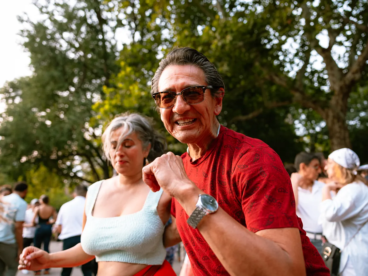 A grey-haired couple dance close together outdoors, the man in a red paisley shirt laughing toward the camera.